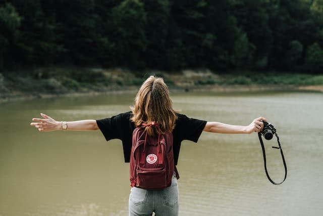 Woman wearing a backpack and standing next to a lake with her arms spread to symbolize overcoming the fear of the unknown