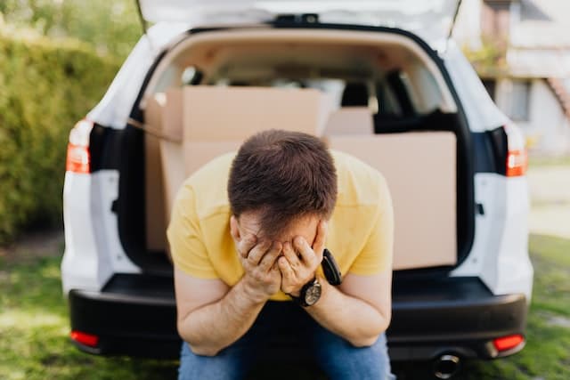 man stressed and sitting on the bumper of car with trunk full of boxes