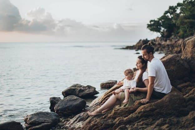 A wife and husband sitting with a baby on a rocky shore.